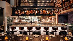 A modern restaurant with a bar area, dining tables set with candles, and a kitchen in the background. - Hotel EMC2, Autograph Collection (Chicago)