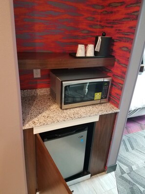 Una kitchenette compacta con un microondas sobre una encimera de granito, un refrigerador pequeño debajo y una pared con un estampado rojo.