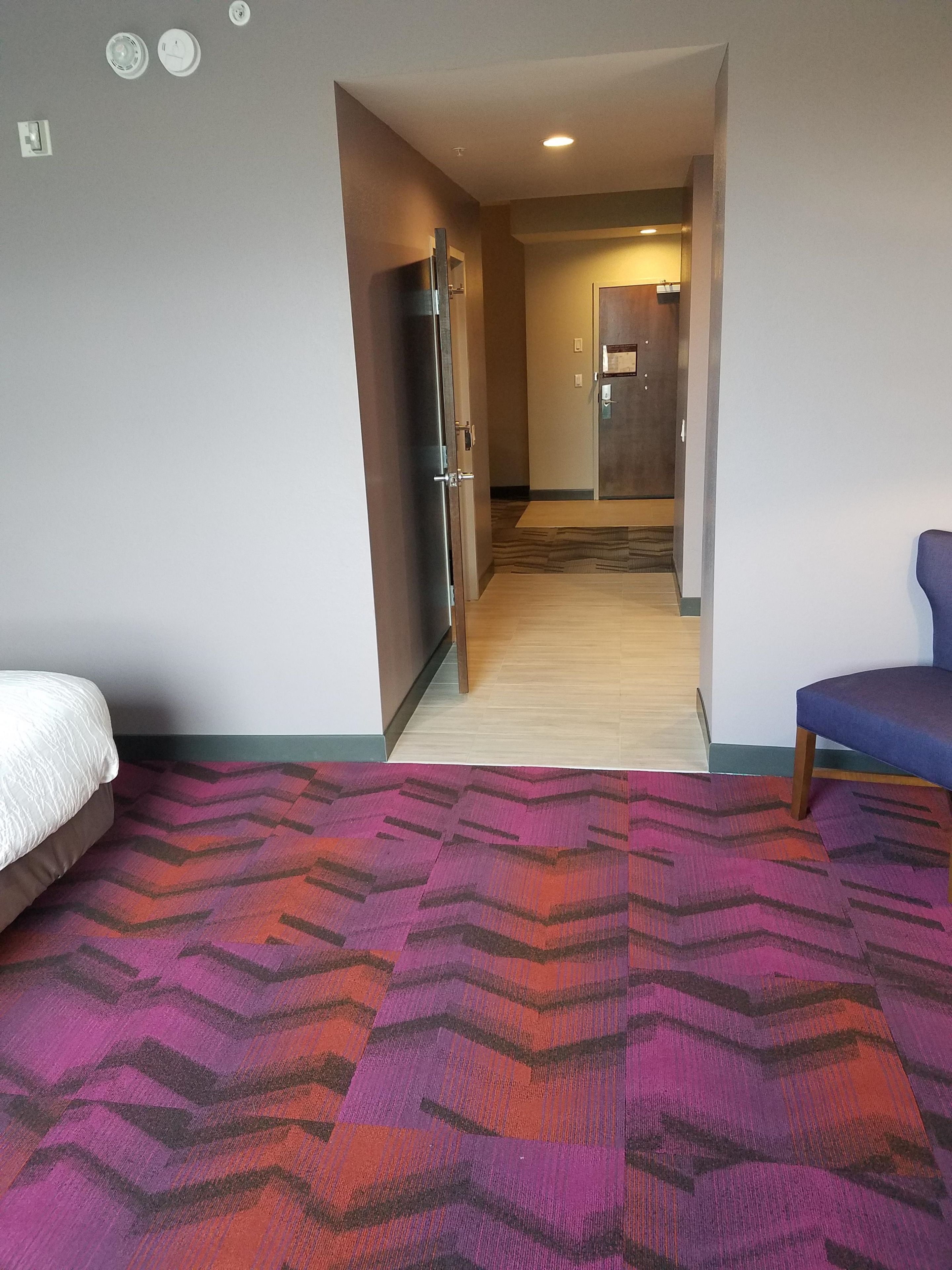 A hotel room with a bed, a blue chair, a patterned rug, a wooden floor, and a hallway leading to a kitchenette.