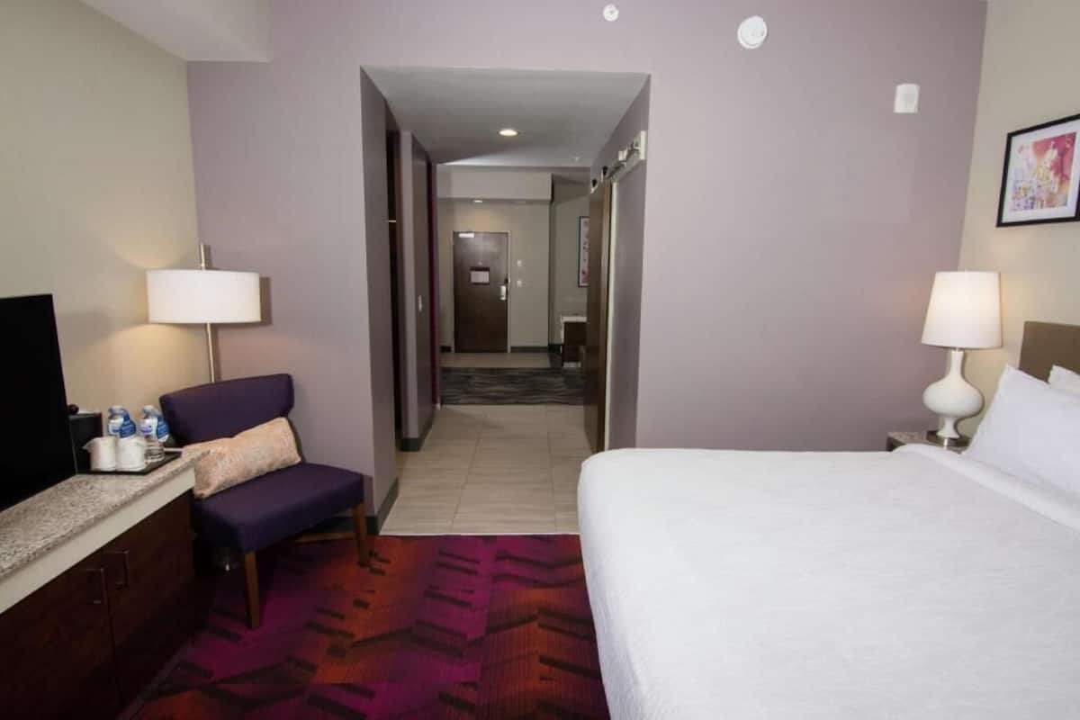 A hotel room with a bed, a desk with a chair, a TV, and a bathroom visible through an open door.