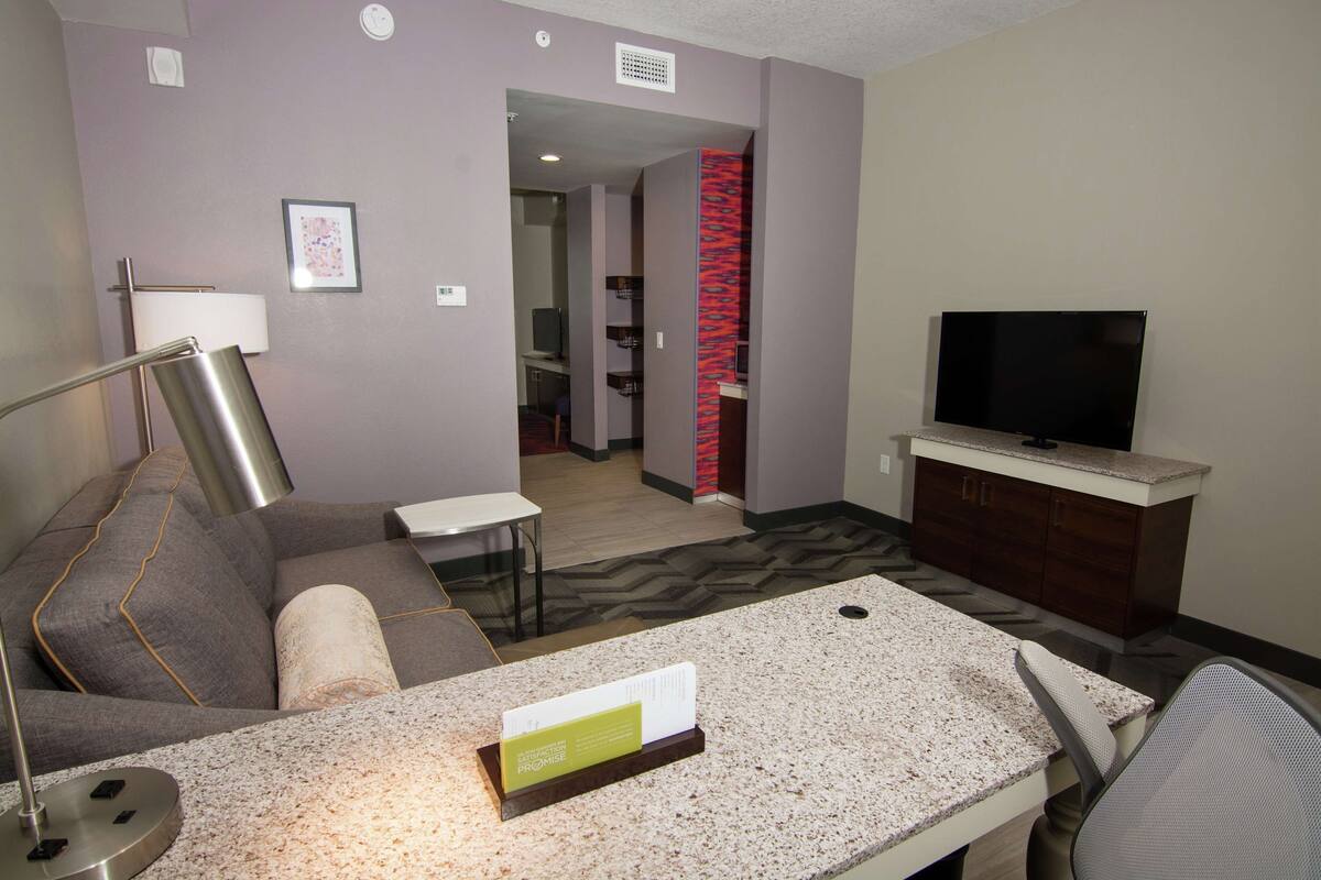 A hotel room with a sofa, a TV, a desk with a sign, and a red accent wall.