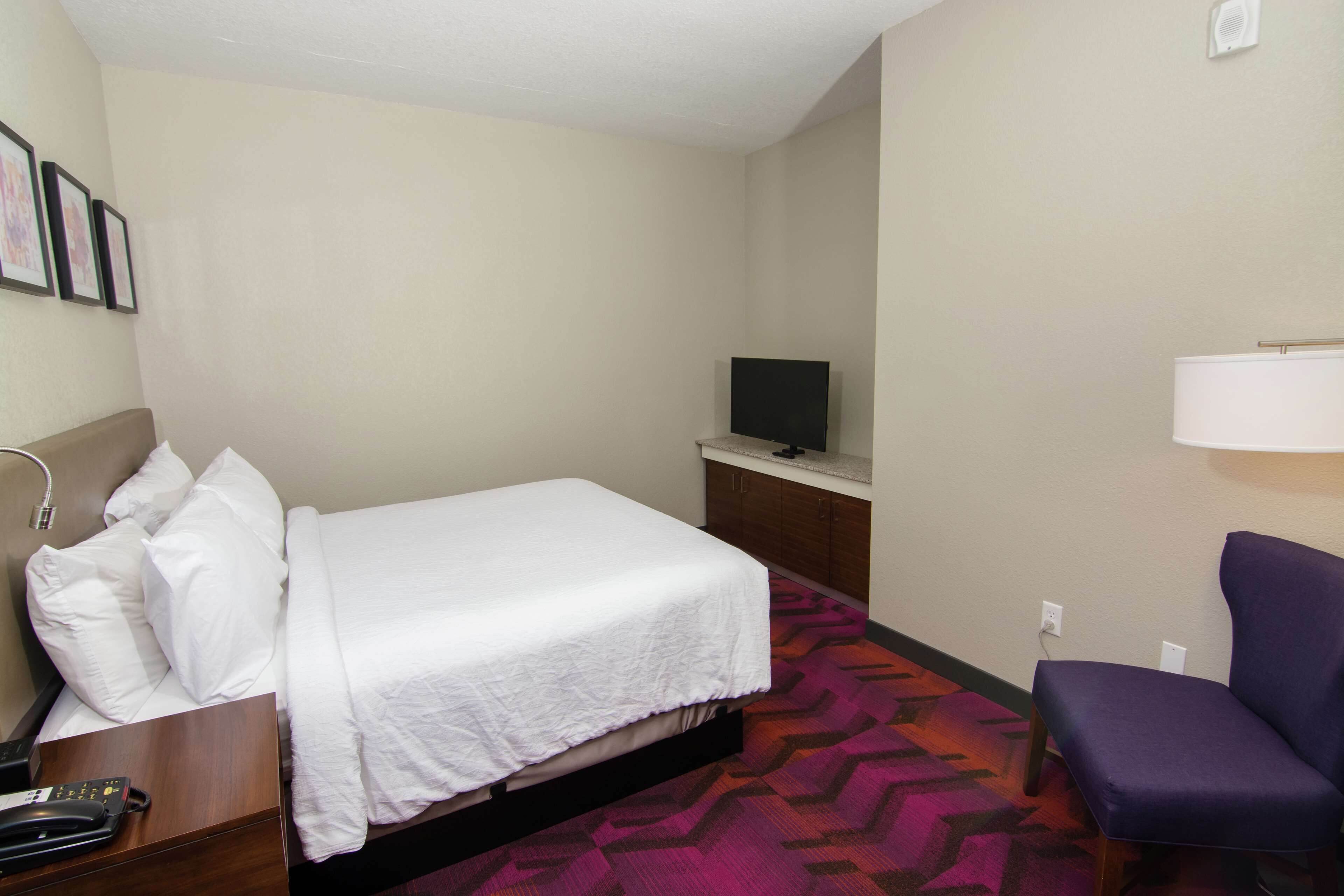 A hotel room with a bed, a television on a stand, a chair, and a desk with a telephone.