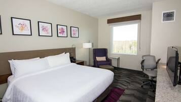A hotel room with a large bed, a desk, a chair, a window, and artwork on the wall.