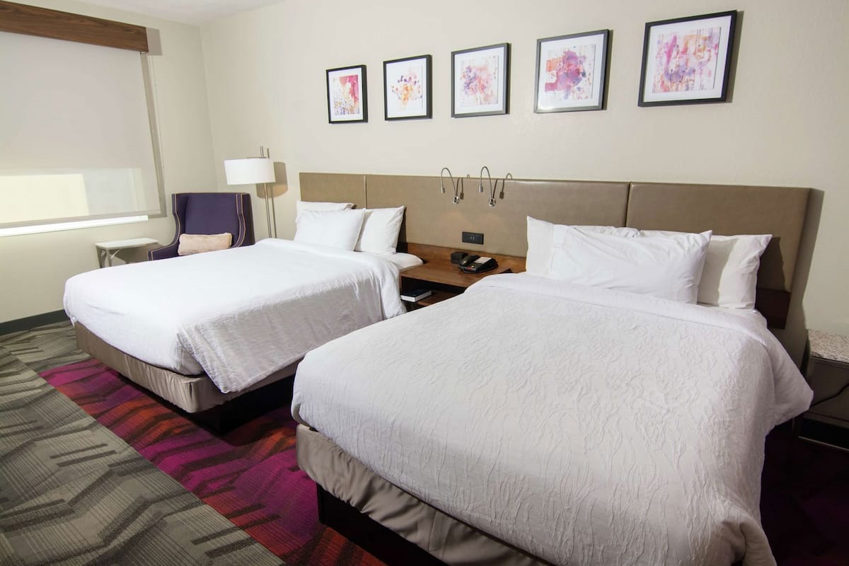 A hotel room with two beds, a nightstand, a lamp, and a window.