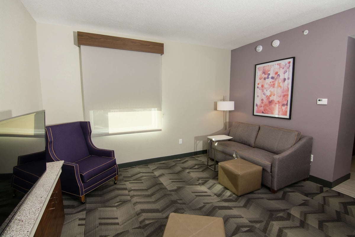 A living room with a purple armchair, a gray sofa, a small table with a lamp, and a large abstract painting on the wall.