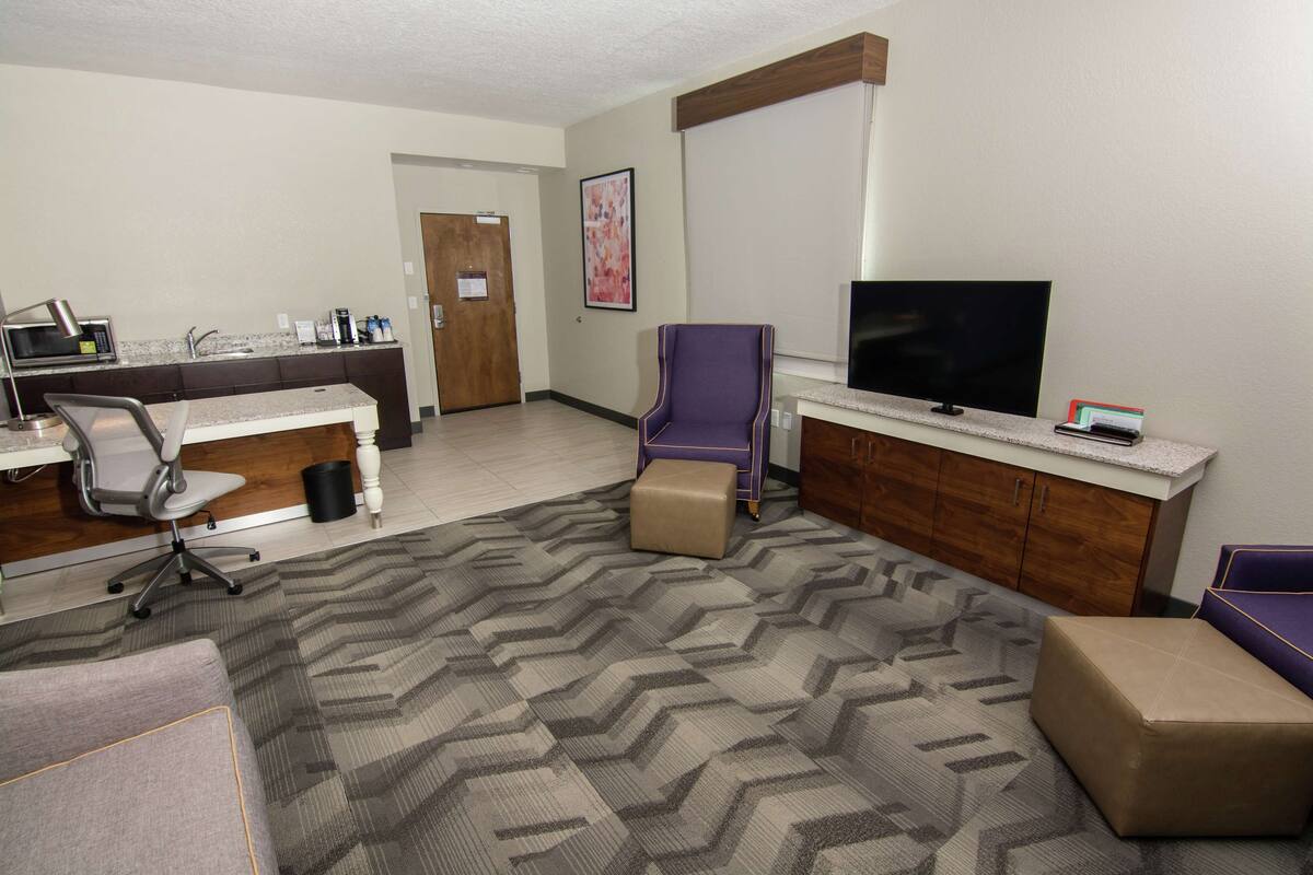 A hotel room with a kitchenette, a TV, a purple armchair, and a sofa.