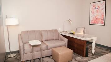 A modern hotel room with a grey sofa, a small table, a desk, a lamp, and a framed abstract painting on the wall.
