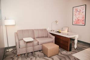 A modern hotel room with a grey sofa, a small table, a desk, a lamp, and a framed abstract painting on the wall. - Hilton Garden Inn Tampa Suncoast Parkway (Lutz)