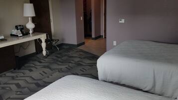 A hotel room with two beds, a desk with a lamp, a coffee maker, and a patterned carpet.
