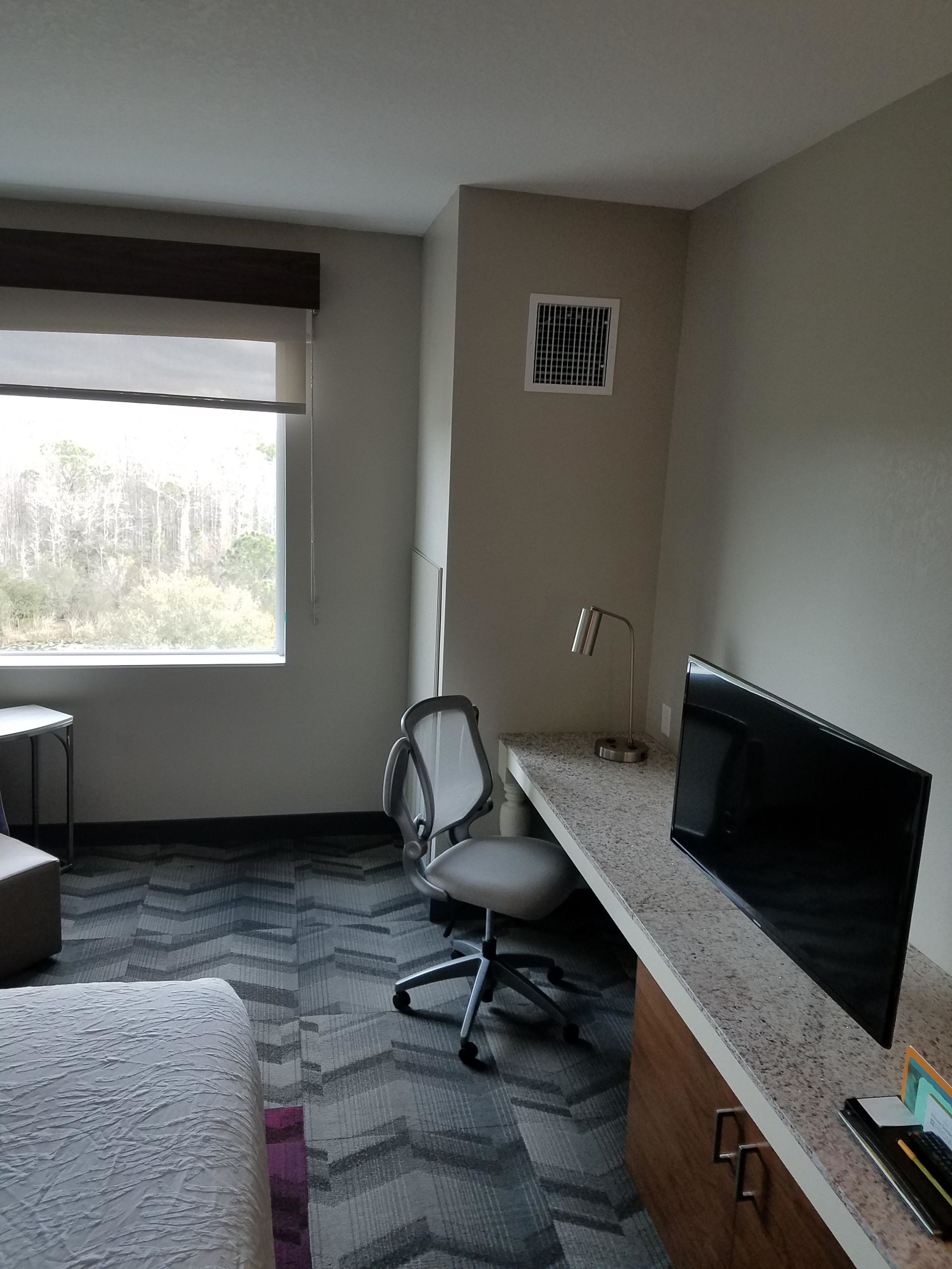 A hotel room with a bed, a desk with a computer monitor, a chair, and a window with a view of trees.