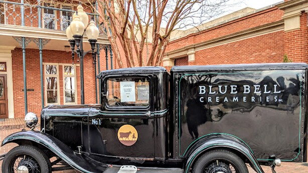 En gammal svart lastbil parkerad framför en byggnad med en fasad av tegelsten, och en skylt som visar texten ”BLUE BELL CREAMERIES”.
