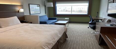 A hotel room with a large bed, a desk with a computer, a chair, a small table, and a window with a view of buildings.