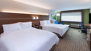 Ett hotellrum med två sängar, ett skrivbord, en stol, ett fönster med persienner och en väggmonterad luftkonditioneringsanläggning.