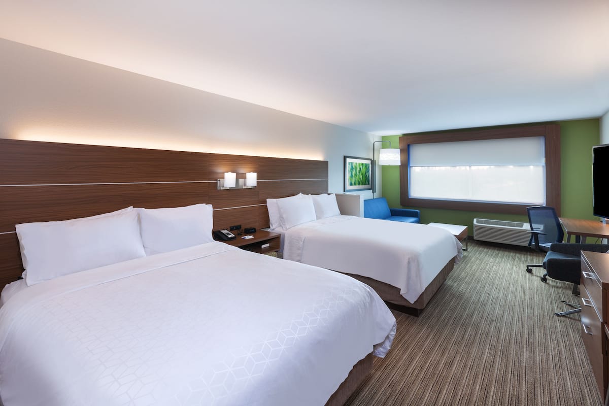 A hotel room with two beds, a desk, a chair, a window with blinds, and a wall-mounted air conditioner.