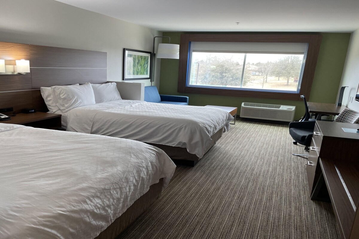 A hotel room with two beds, a desk with a chair, a window with a view, and a wall-mounted lamp.