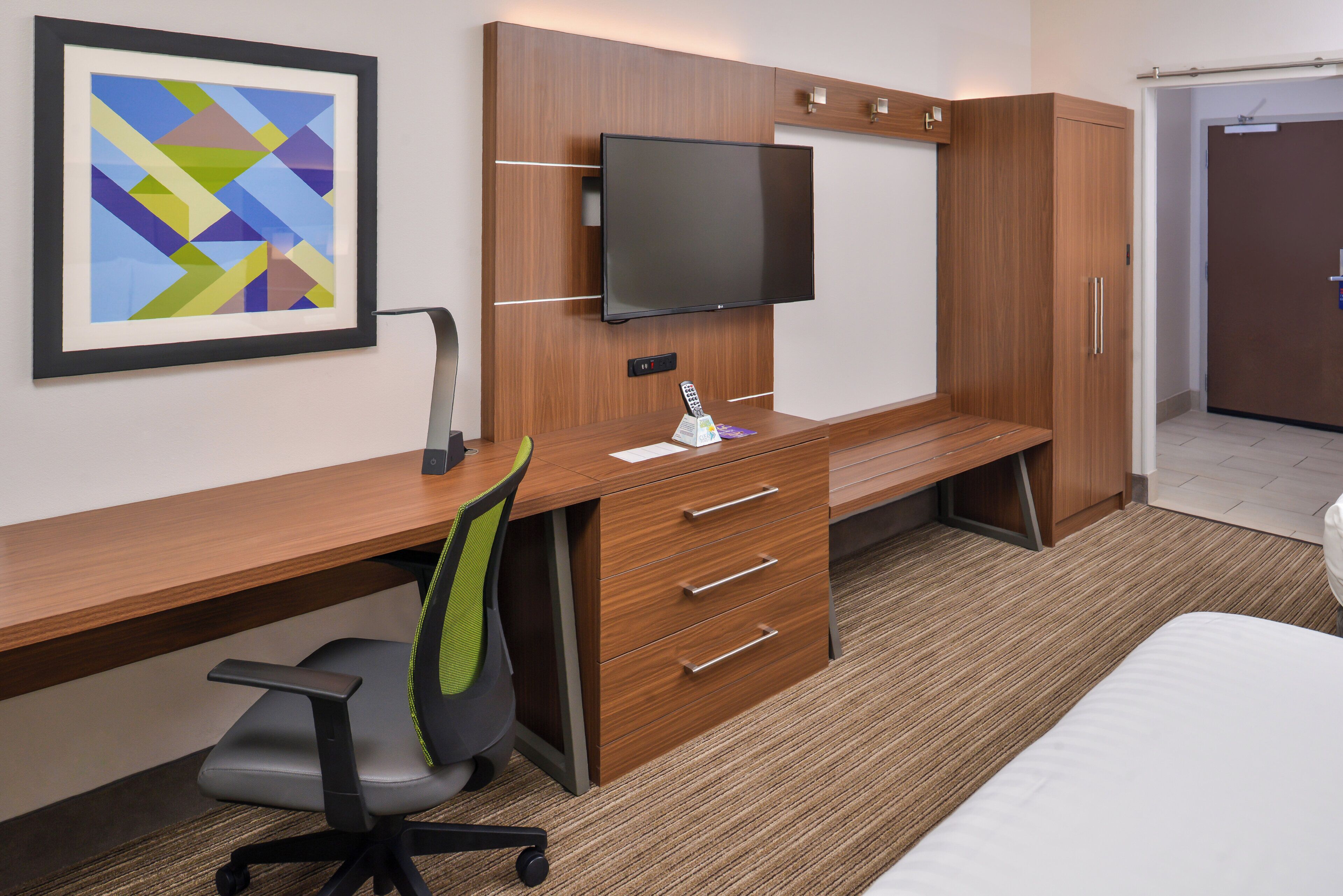 A hotel room with a wooden desk, a flat-screen TV, a chair, a bed, and a framed artwork on the wall.