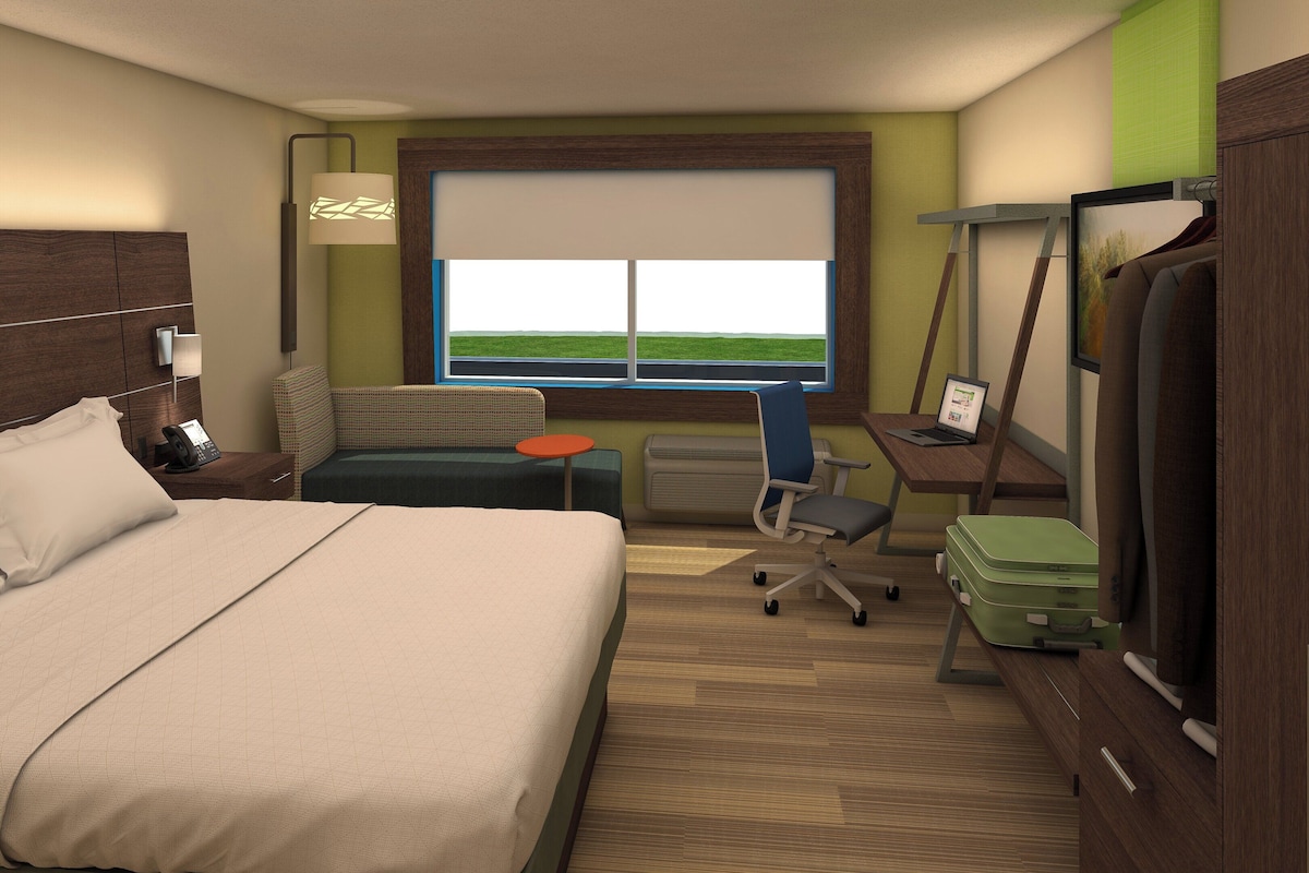 A hotel room with a bed, a desk with a laptop, a chair, a small table, a suitcase, and a window with a view.