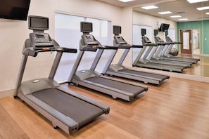 A row of stationary treadmills in a gym with a mirrored wall and a television screen above each machine.