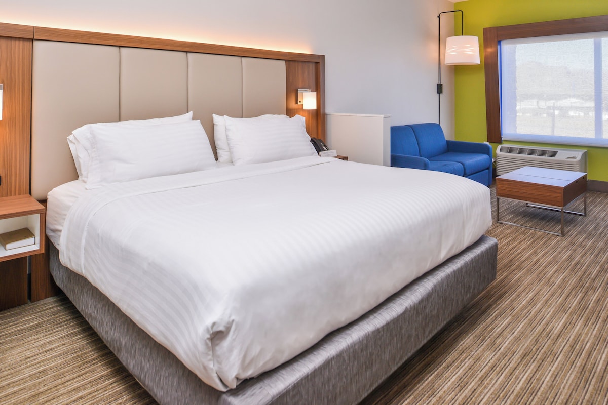 A hotel room with a large bed, a desk, a chair, a window, and a wall-mounted lamp.
