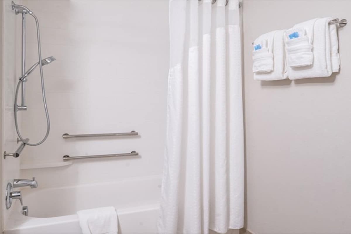 Un baño con bañera, ducha con cabezal de ducha manual, barras de apoyo y un perchero con toallas.