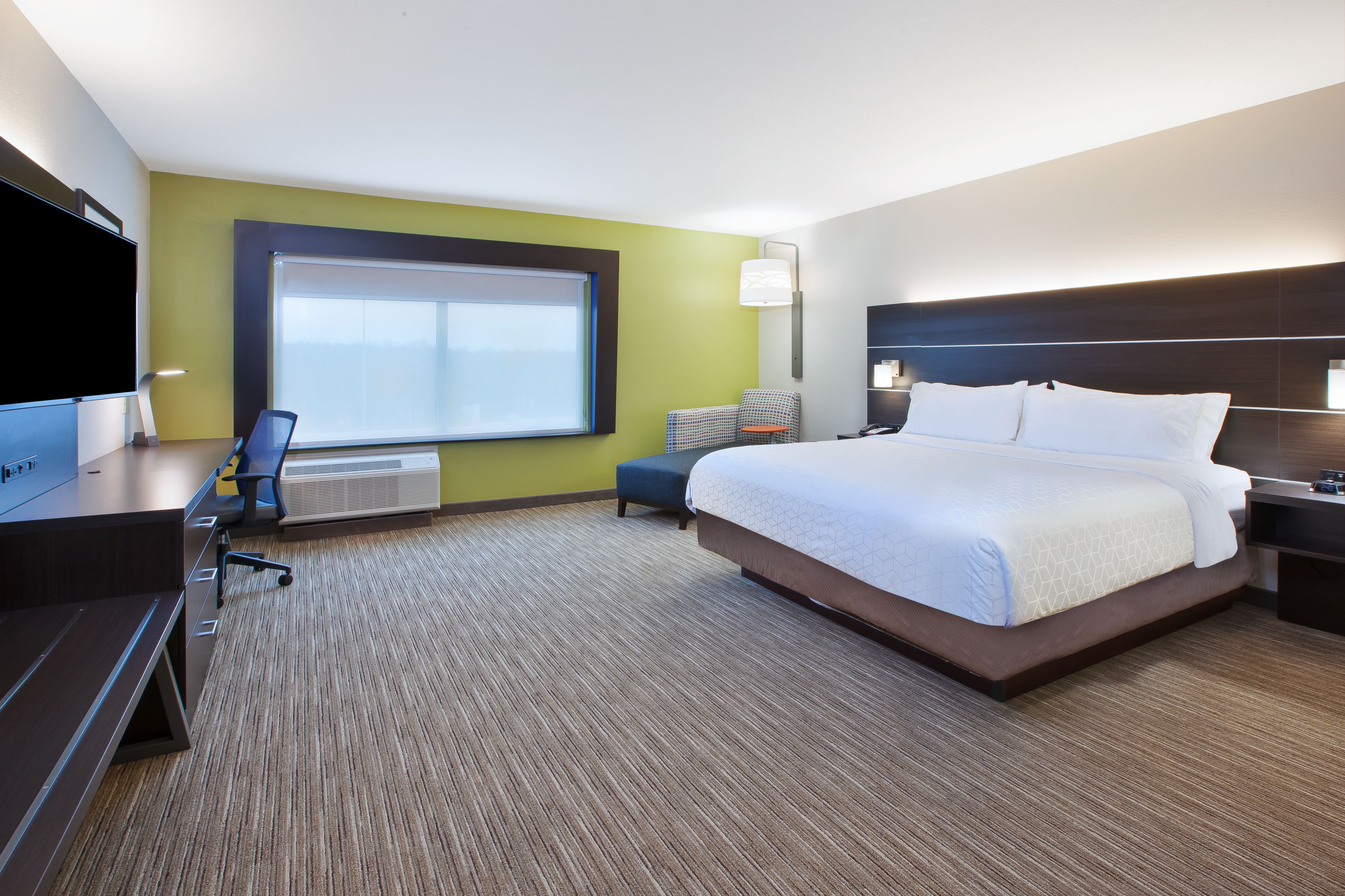 Une chambre d’hôtel avec un grand lit, un bureau avec une chaise, une télévision et une fenêtre avec des stores.
