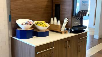 Estación autoservicio de frutas con un bol de fruta, una máquina de café y un letrero que dice “Consiéntete”.