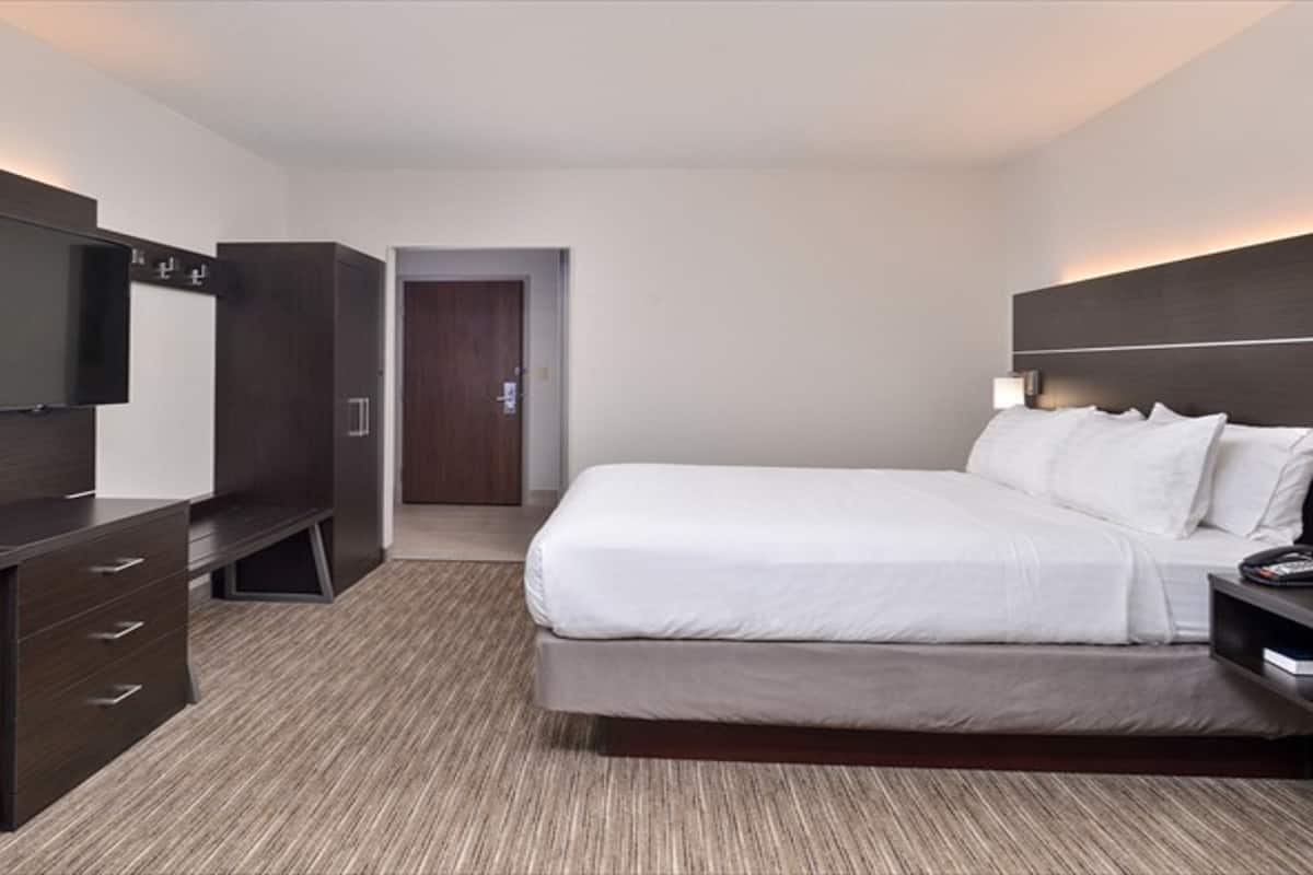 A modern hotel room with a large bed, a TV mounted on the wall, a desk with drawers, and a door leading to another room.