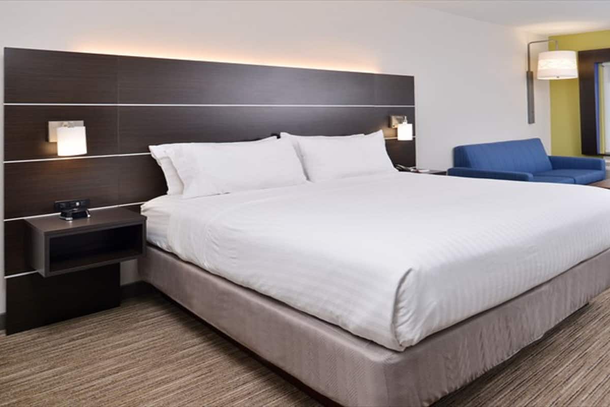 A hotel room with a large bed, a nightstand, a desk, a blue chair, and a large window.