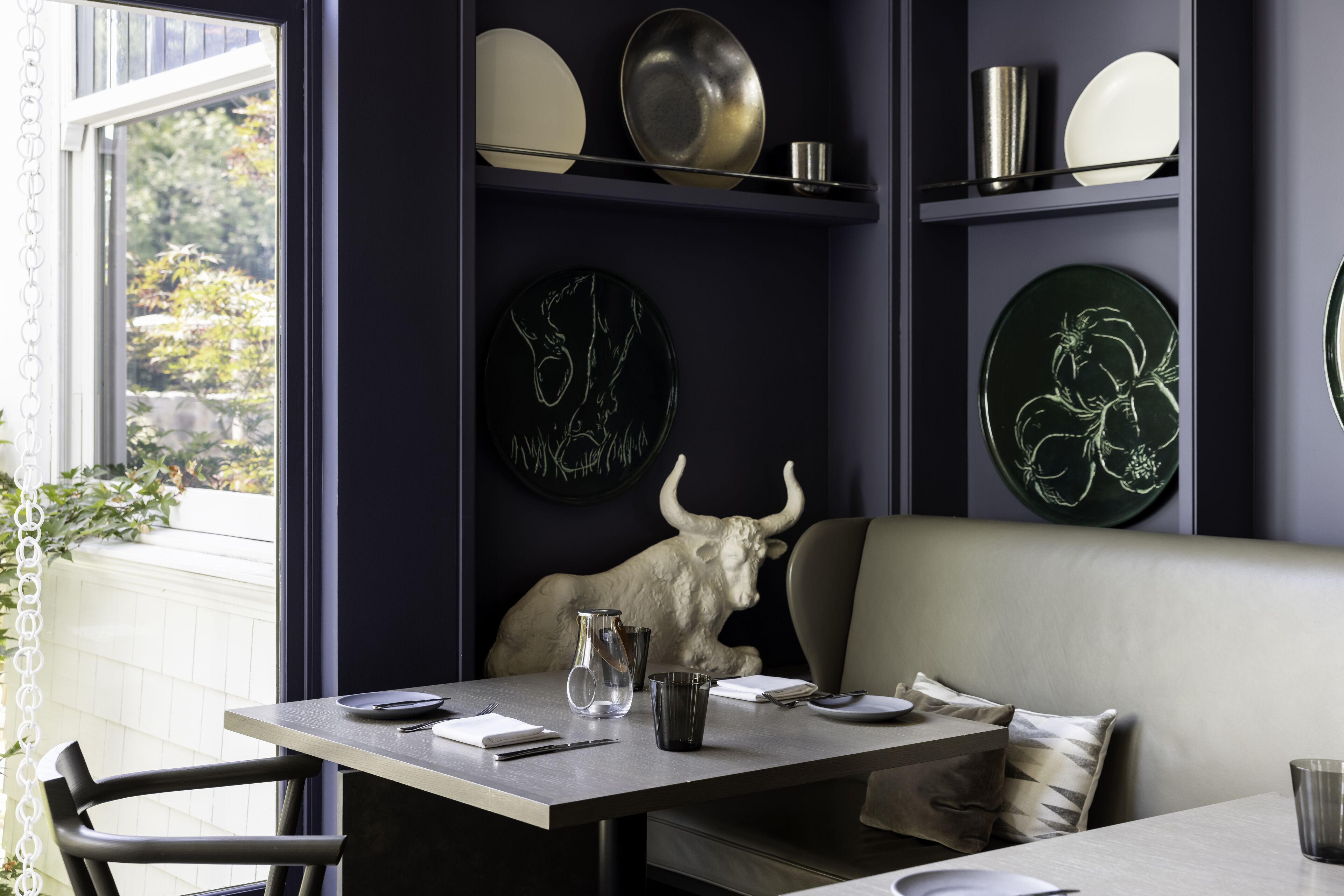 A dining area with a table set for two, a bull sculpture, and decorative plates on shelves.