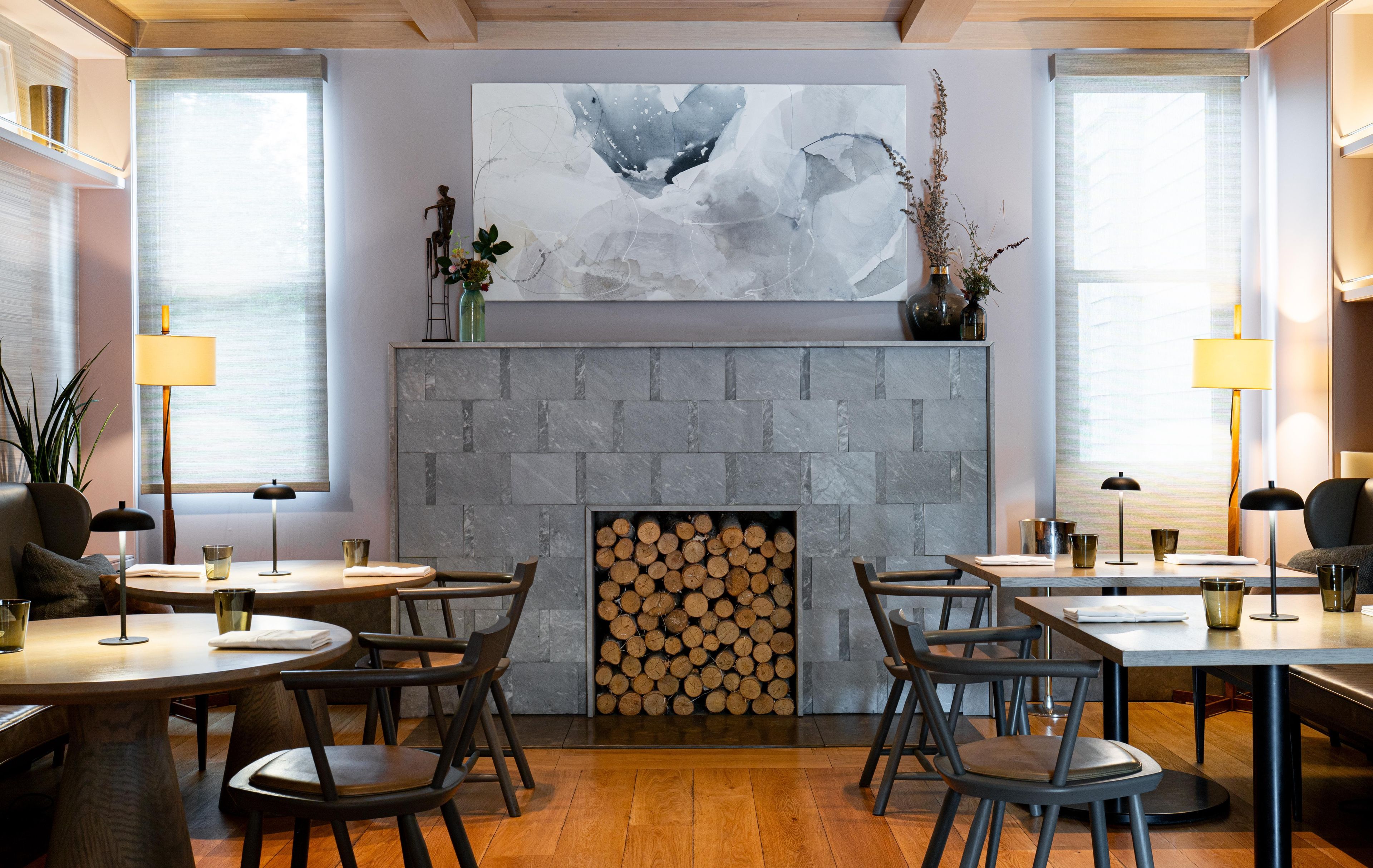 Un comedor moderno con mesas redondas, sillas, una chimenea con leña apilada y arte abstracto en la pared.