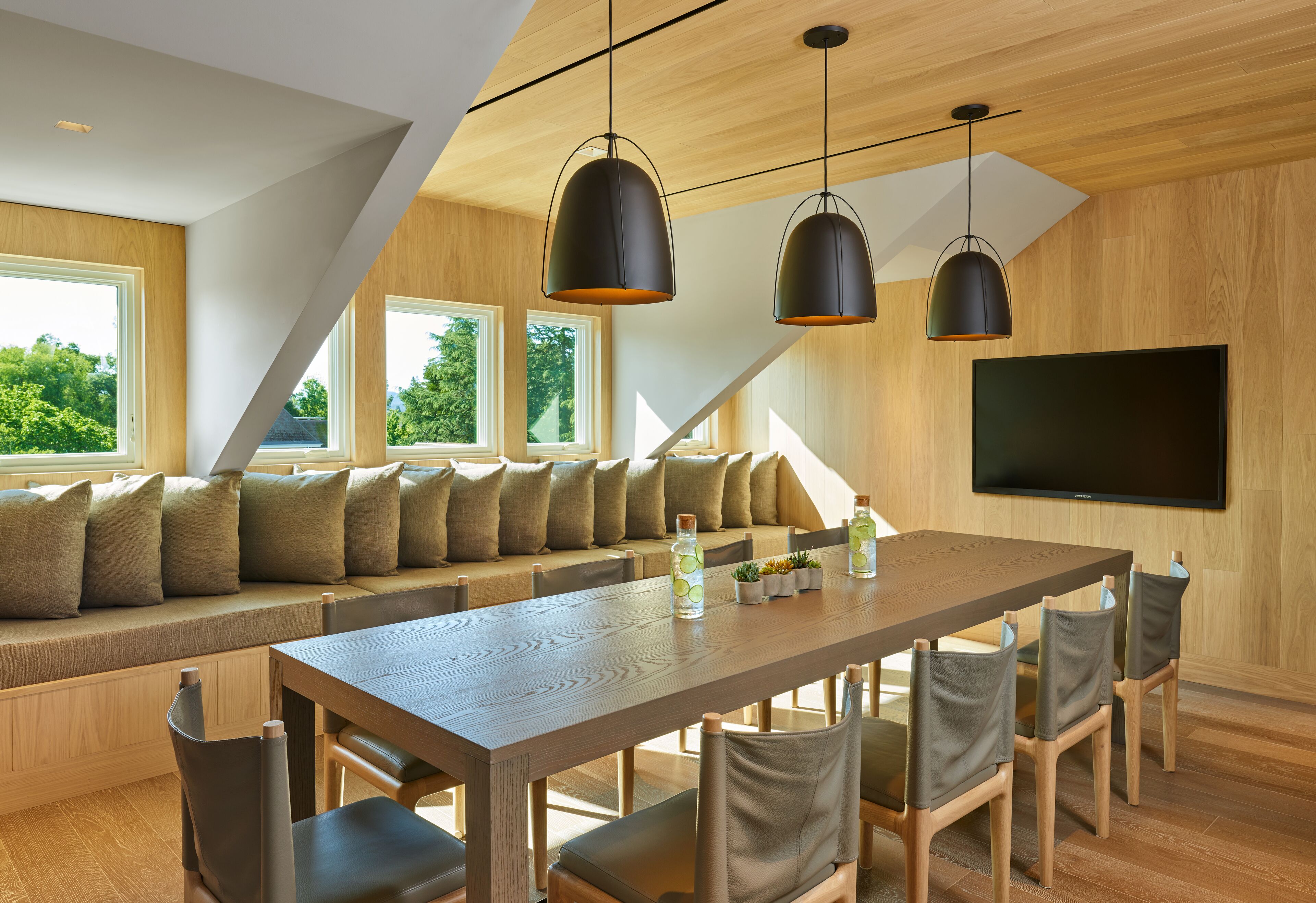 A modern dining area with a long wooden table, cushioned seating, pendant lighting, and a flat-screen TV on the wall.