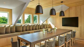 A modern dining area with a long wooden table, cushioned seating, pendant lighting, and a flat-screen TV on the wall.
