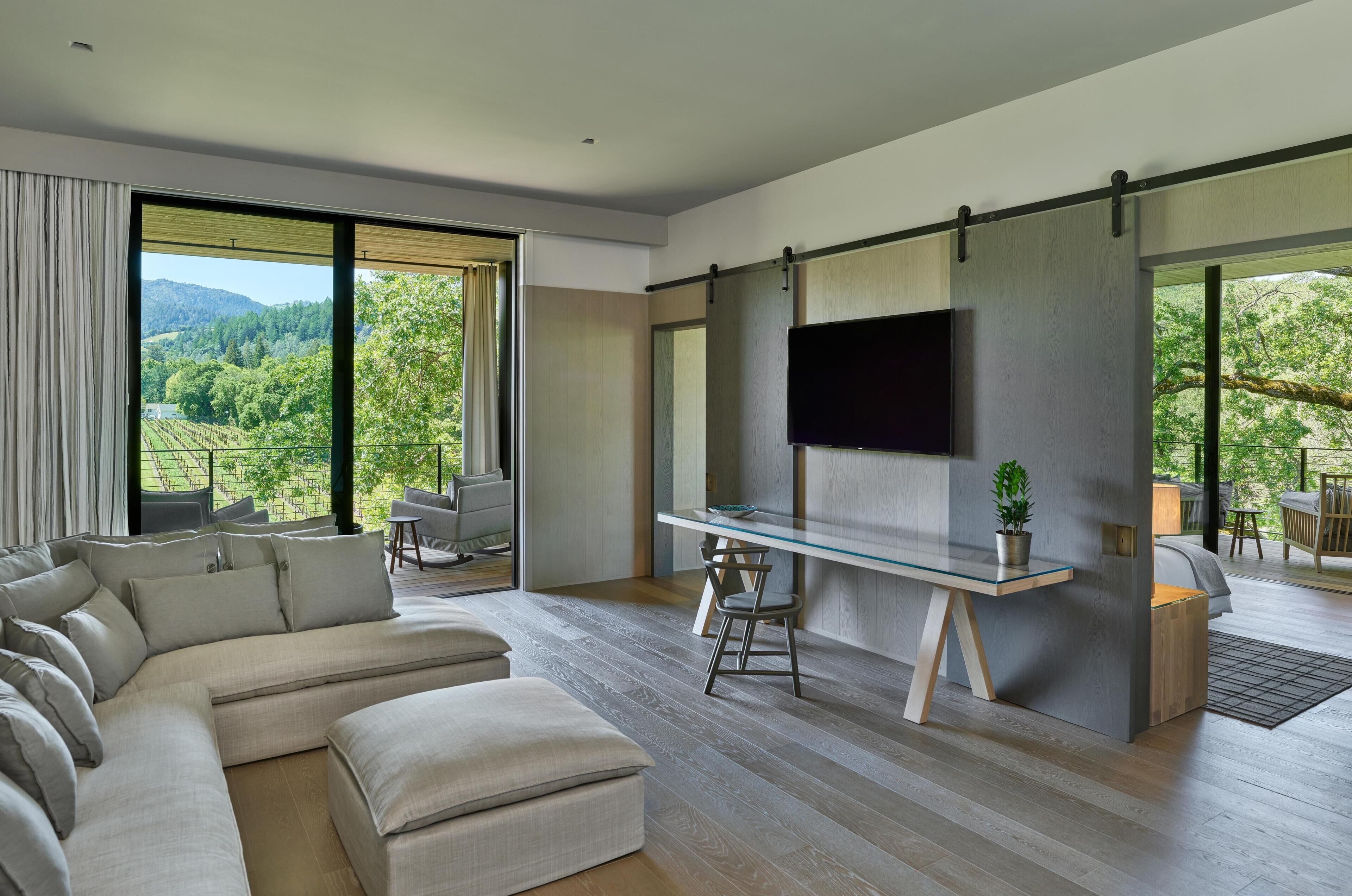 A modern living room with a large sectional sofa, a glass dining table, and a flat-screen TV mounted on a wooden wall.