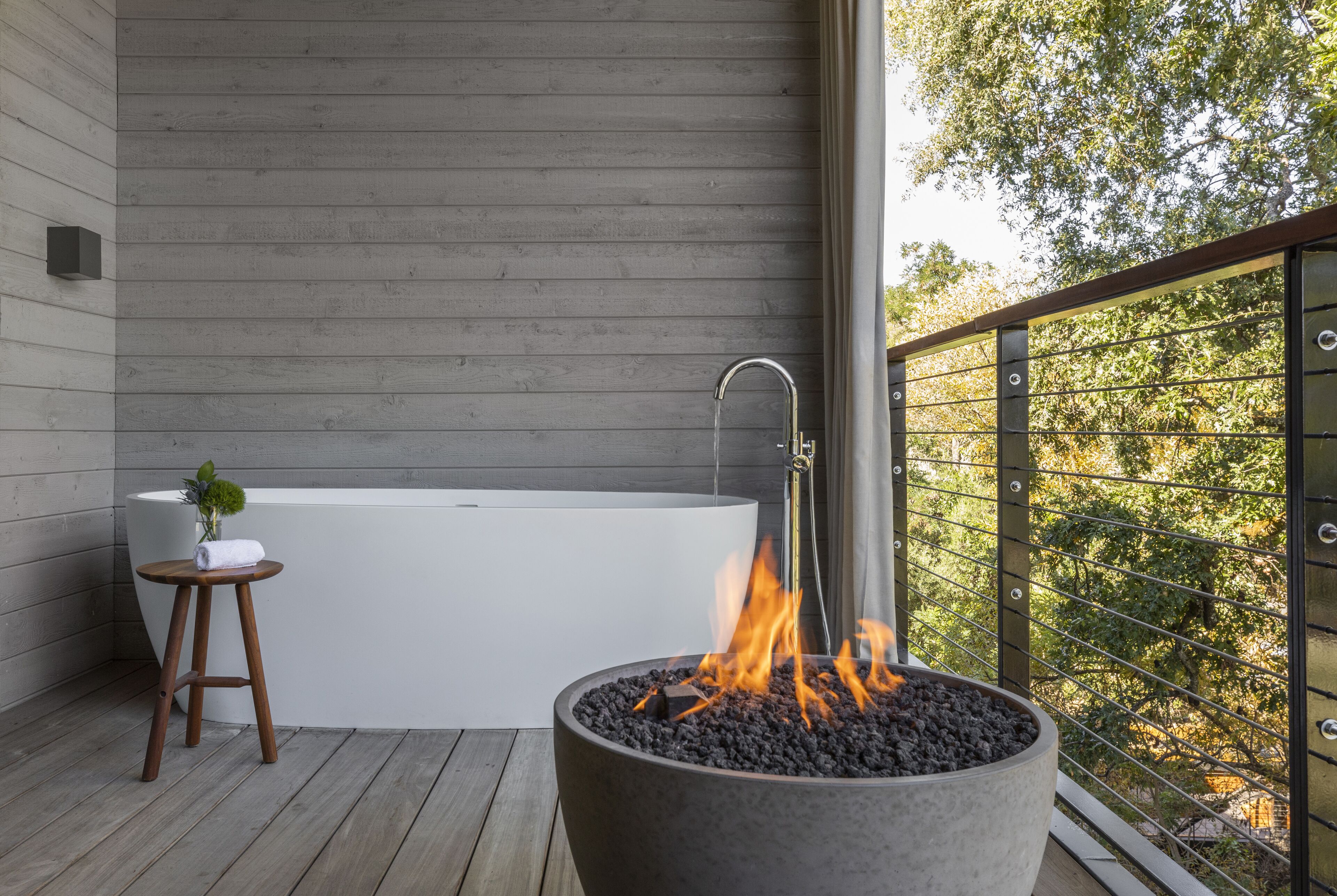 A modern outdoor bathroom with a bathtub, a fire pit, and a wooden stool.