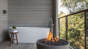 A modern outdoor bathroom with a bathtub, a fire pit, and a wooden stool.