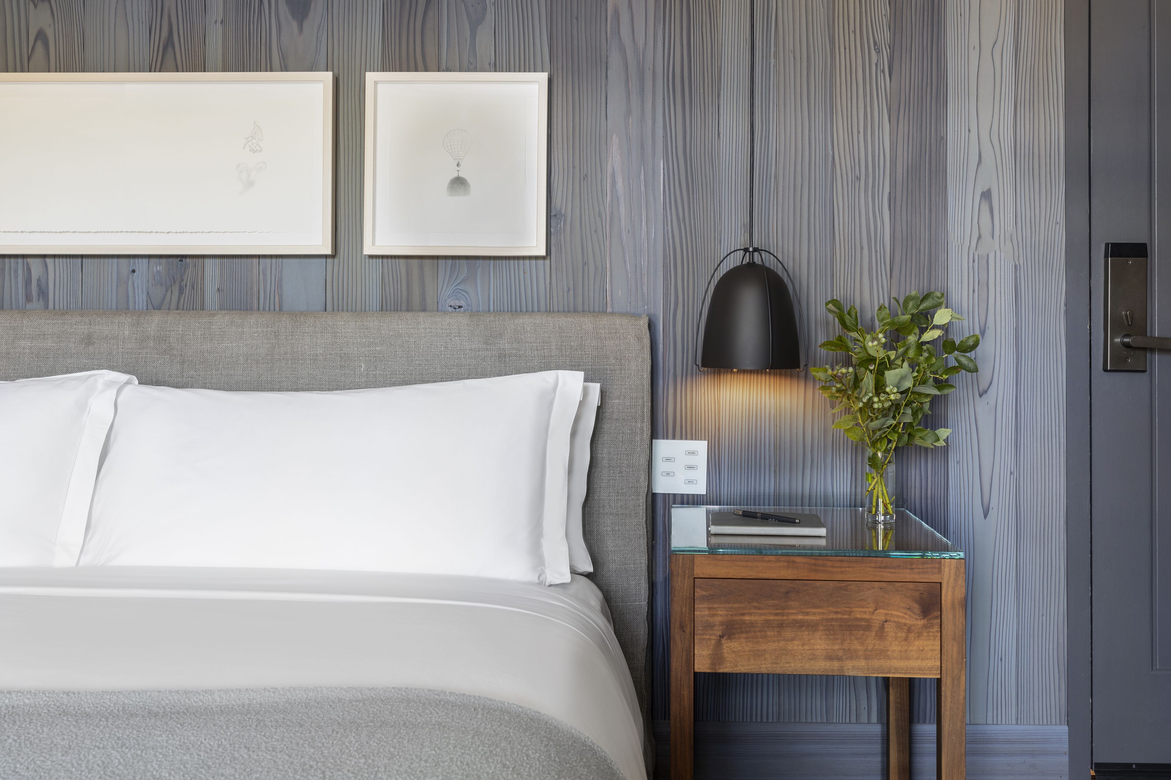 Een net opgemaakt bed met witte lakens, een nachtkastje met glazen blad, een zwarte lamp en een kleine potplant.