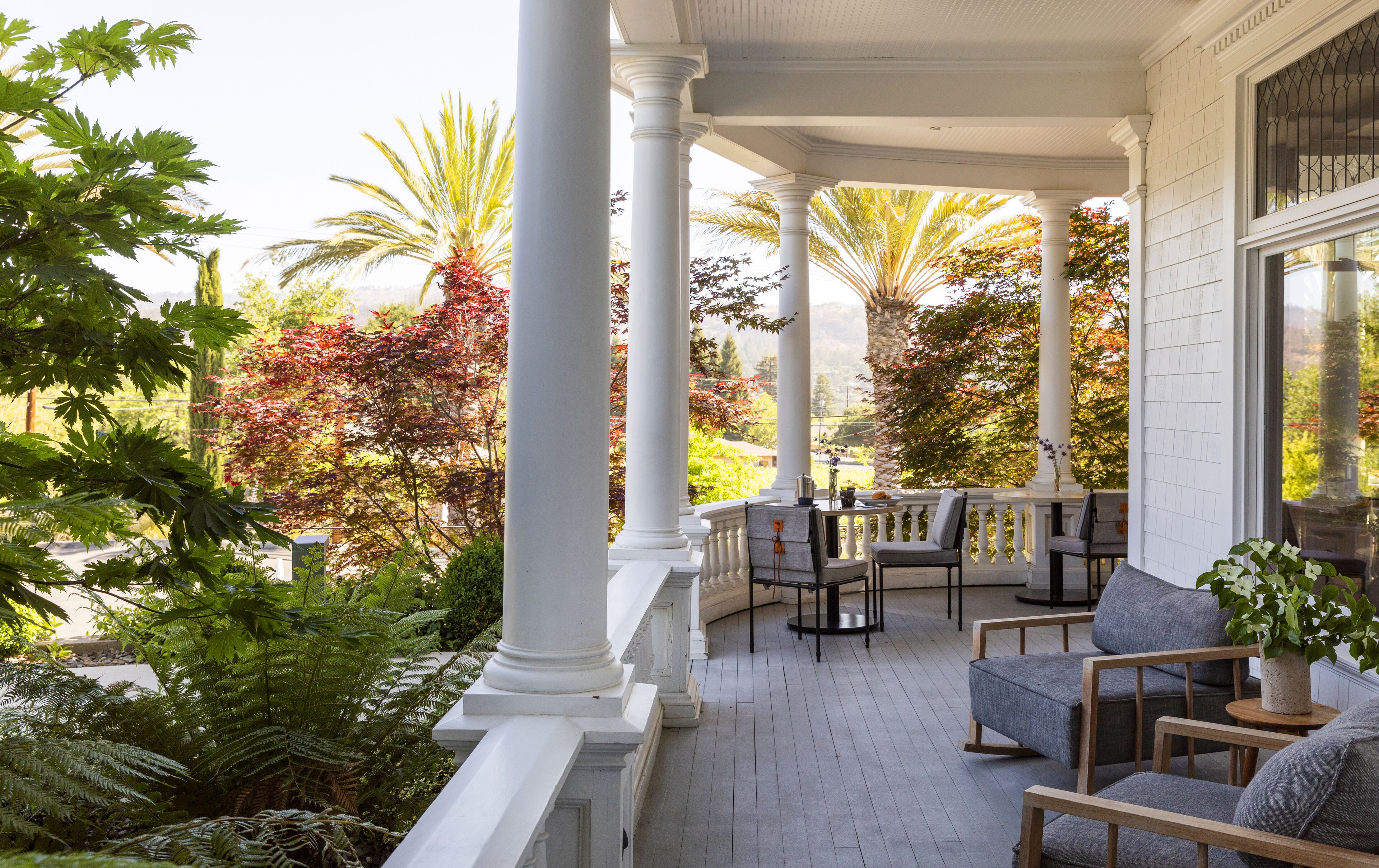 A covered porch with white columns, outdoor seating, and a view of a garden with various trees.