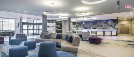 A spacious lobby with a large curved sofa, multiple ottomans, a reception desk, and a modern geometric wall design.