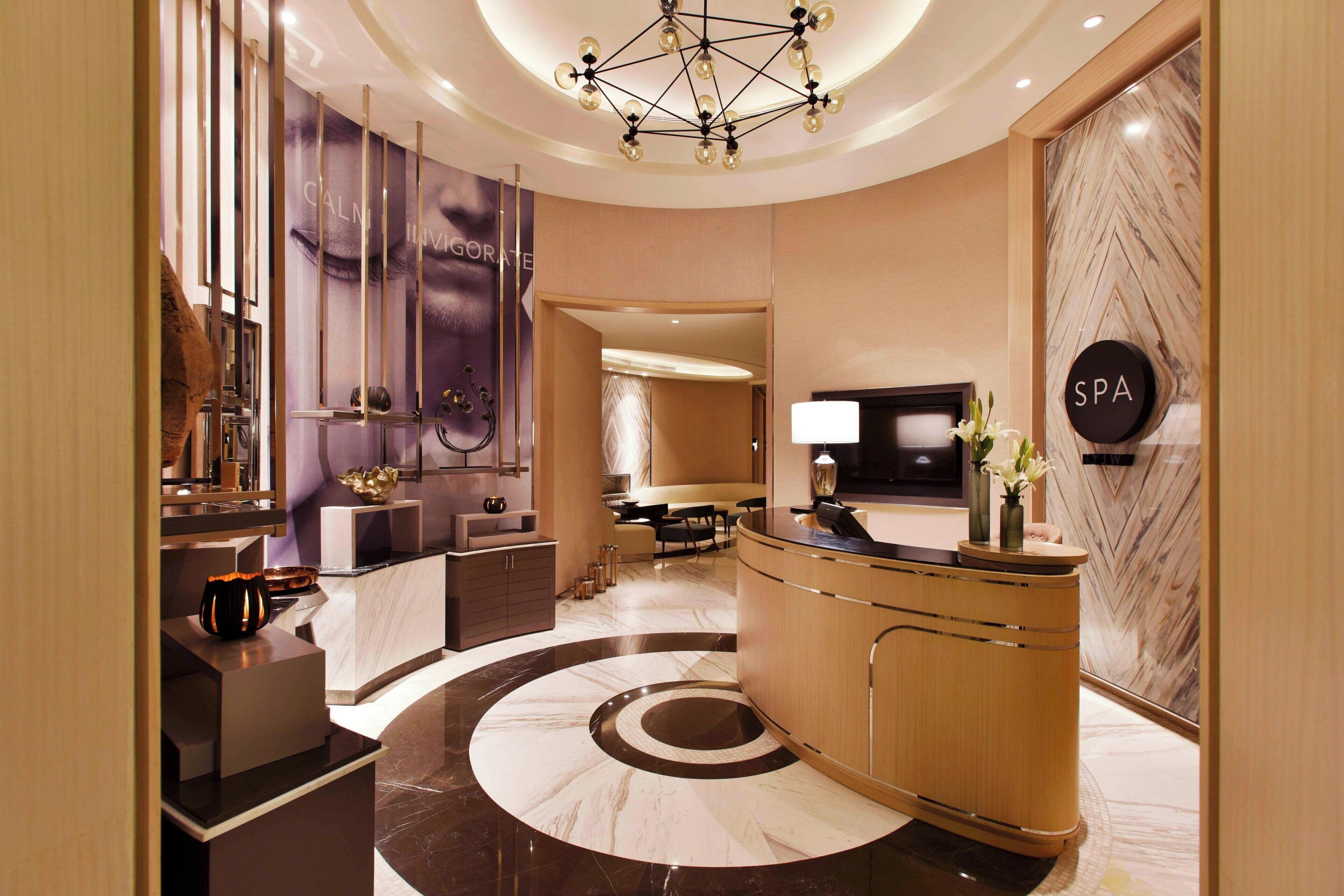 Um lobby de hotel moderno com balcão de recepção, placa indicando spa e um espelho grande refletindo o interior.