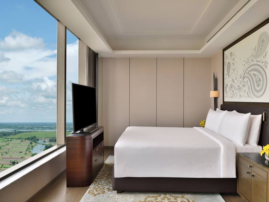 A modern hotel room with a large bed, a flat-screen TV, a nightstand, and a view of a landscape through a large window.