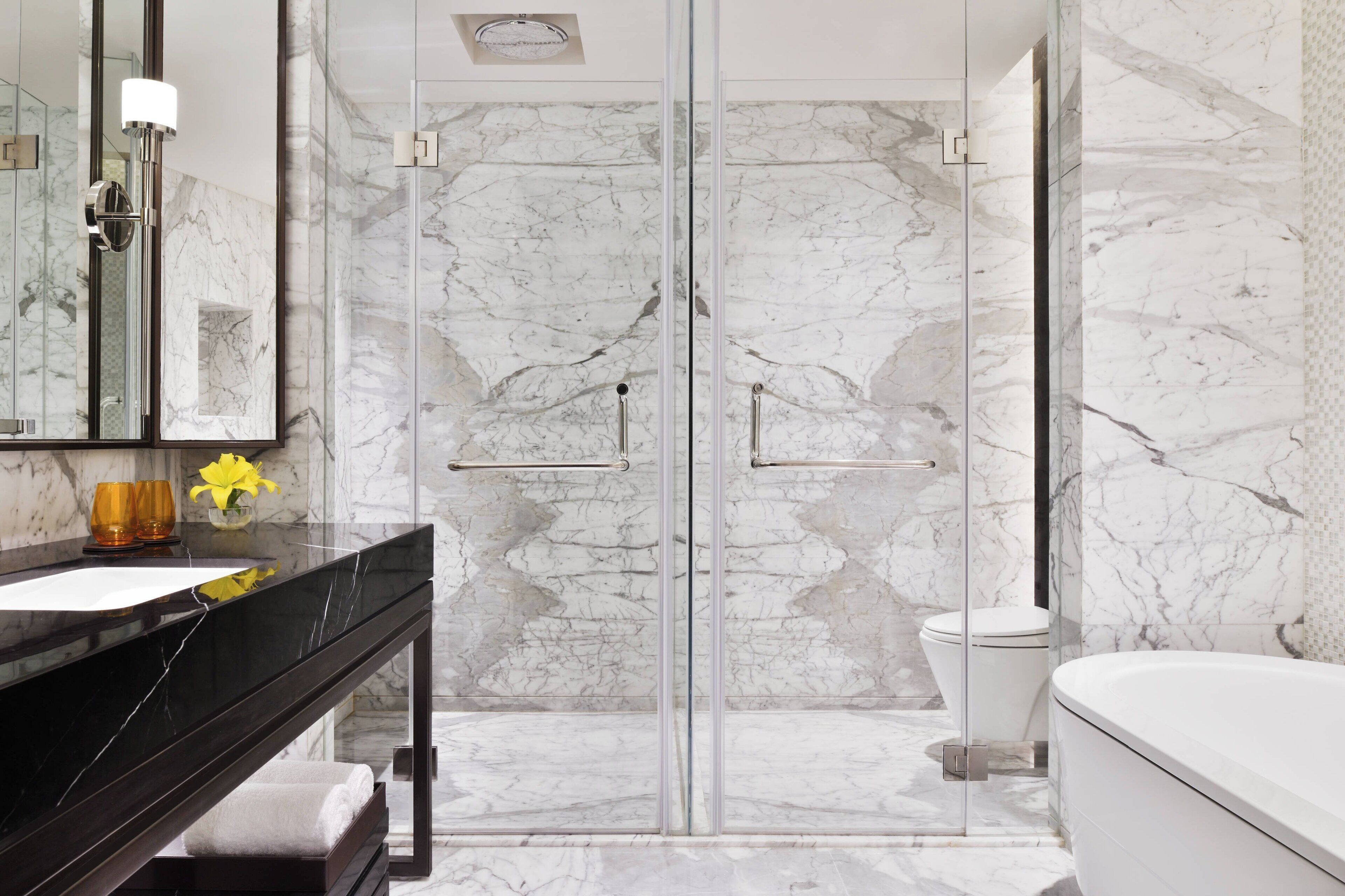 A modern bathroom with a glass-enclosed shower, a marble-tiled wall, a black countertop, and a white bathtub.