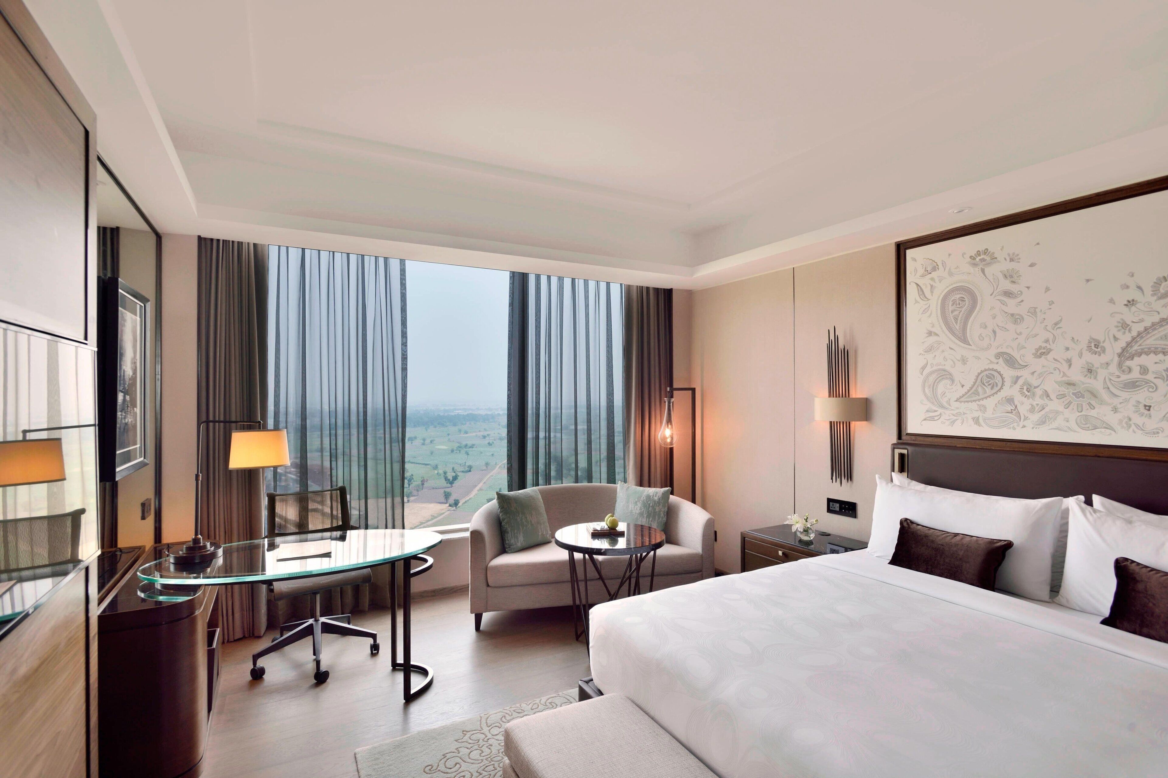 A hotel room with a large bed, a desk with a chair, a small table, and a sofa. There is a large window with curtains, a wall-mounted lamp, and a framed artwork above the bed.
