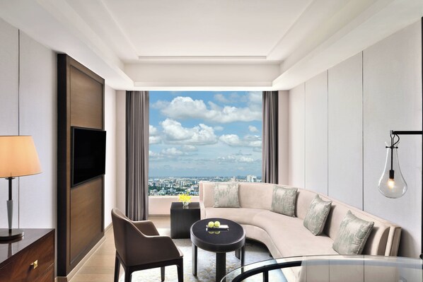 Habitación de hotel moderna con un sofá curvo, una pequeña mesa redonda, un televisor montado en la pared y un ventanal con vistas a la ciudad.