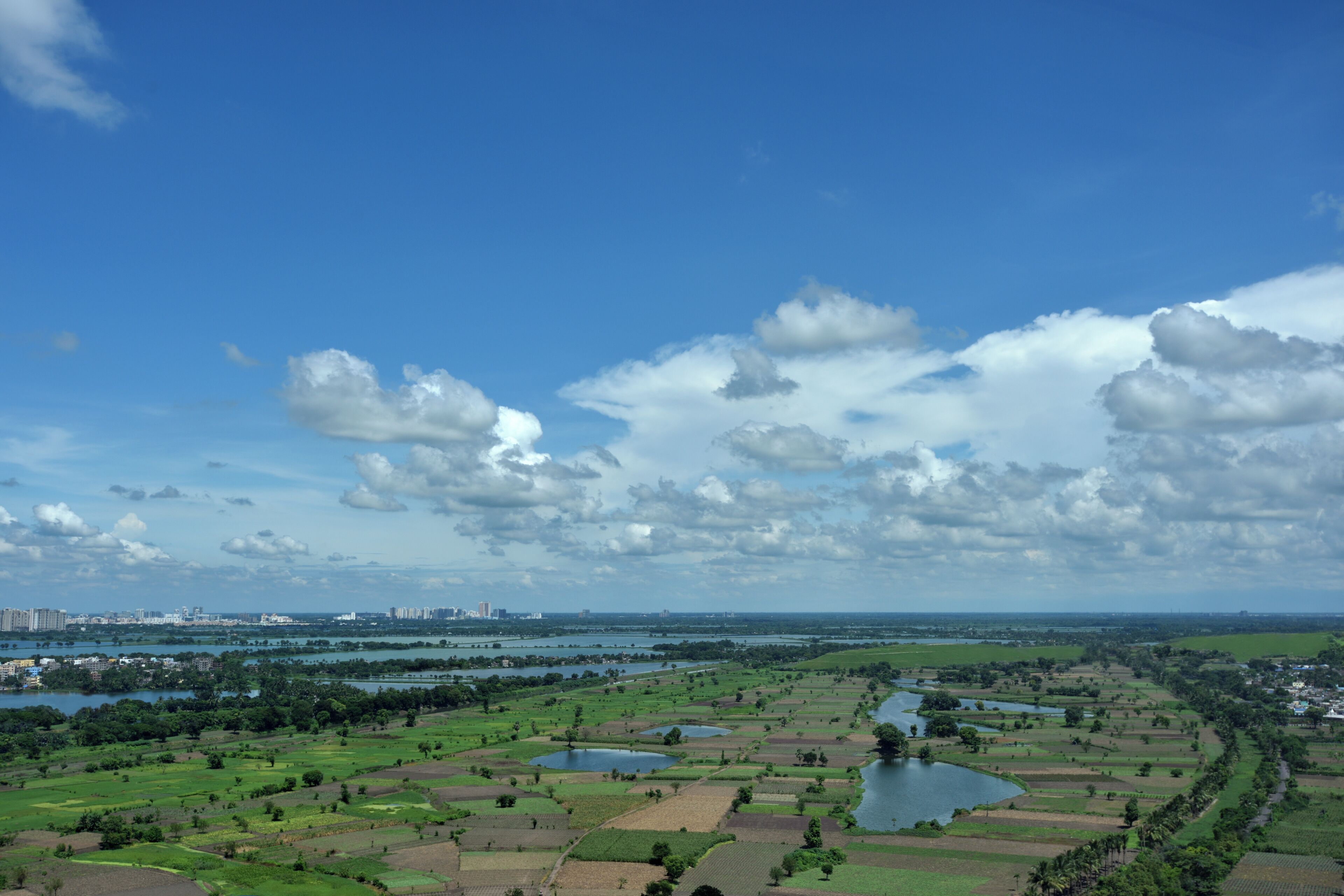 A panoramic view of a landscape with fields, water bodies, and distant buildings.