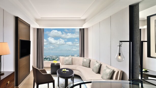 A modern hotel room with a curved sofa, a small round table, a TV, and a large window offering a city view.
