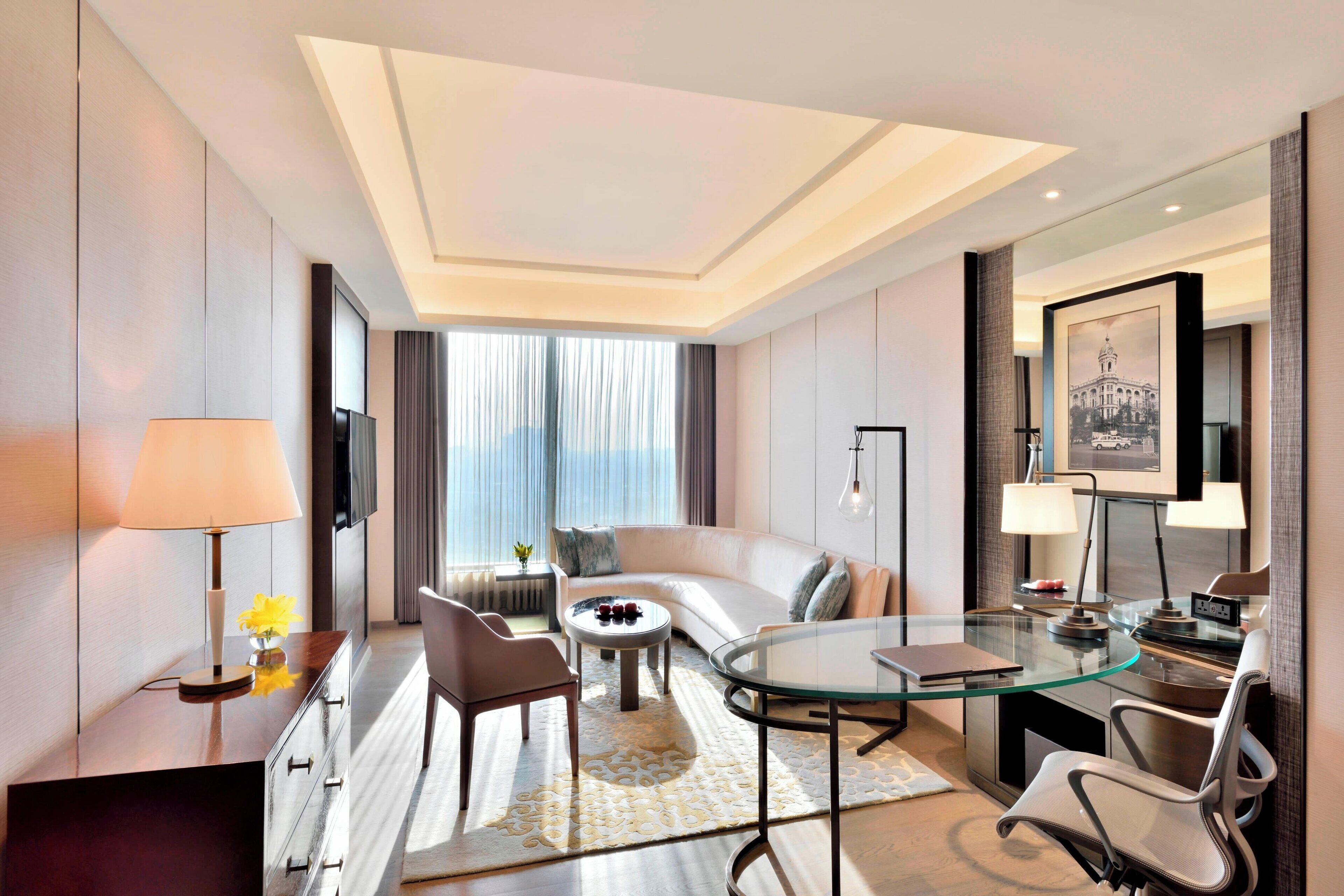 A modern hotel room with a curved sofa, a glass coffee table, a desk with a chair, and a large window with curtains.