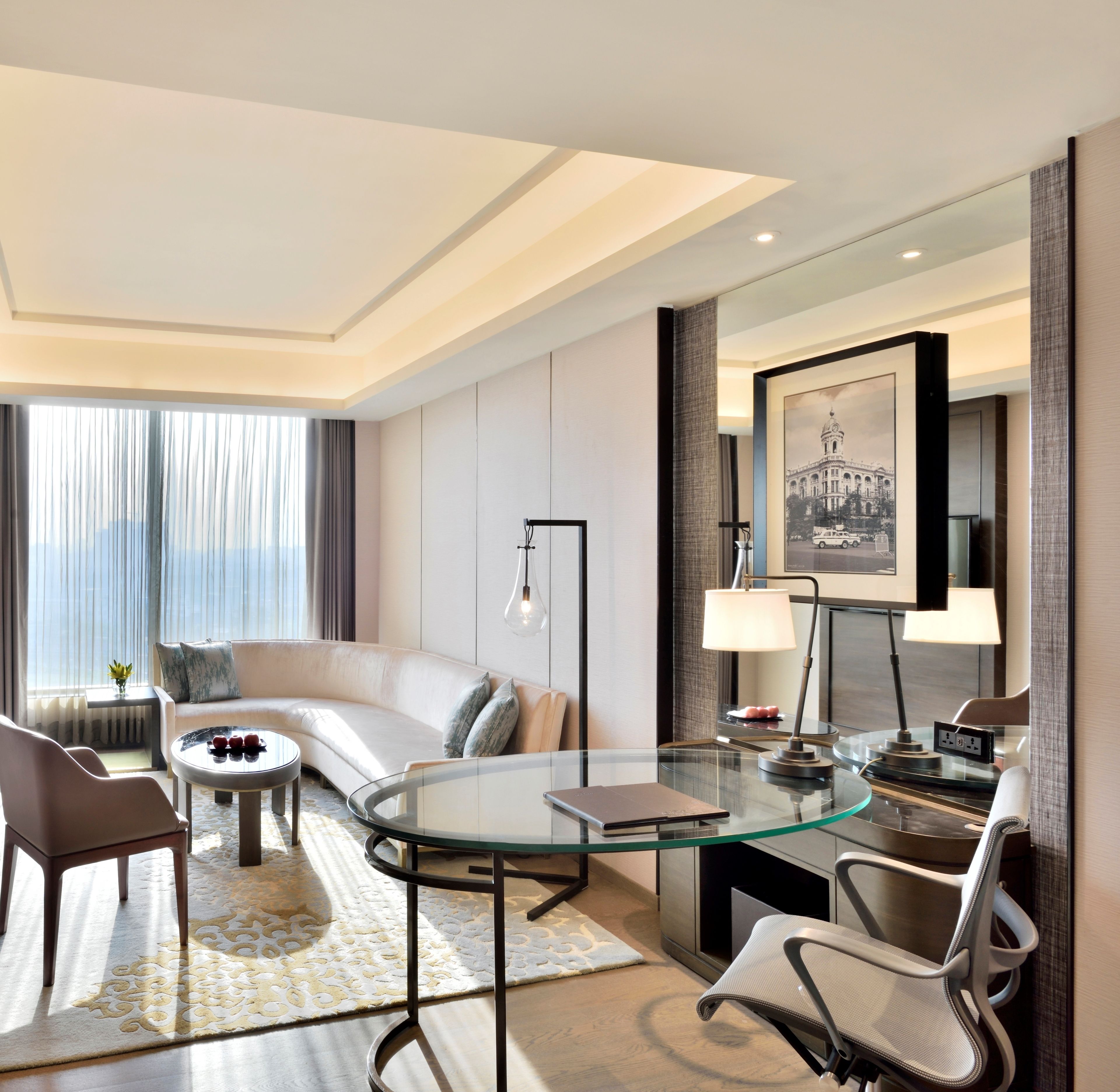 Habitació d'hotel moderna amb taula de vidre, un sofà, una cadira i escriptori amb làmpada.