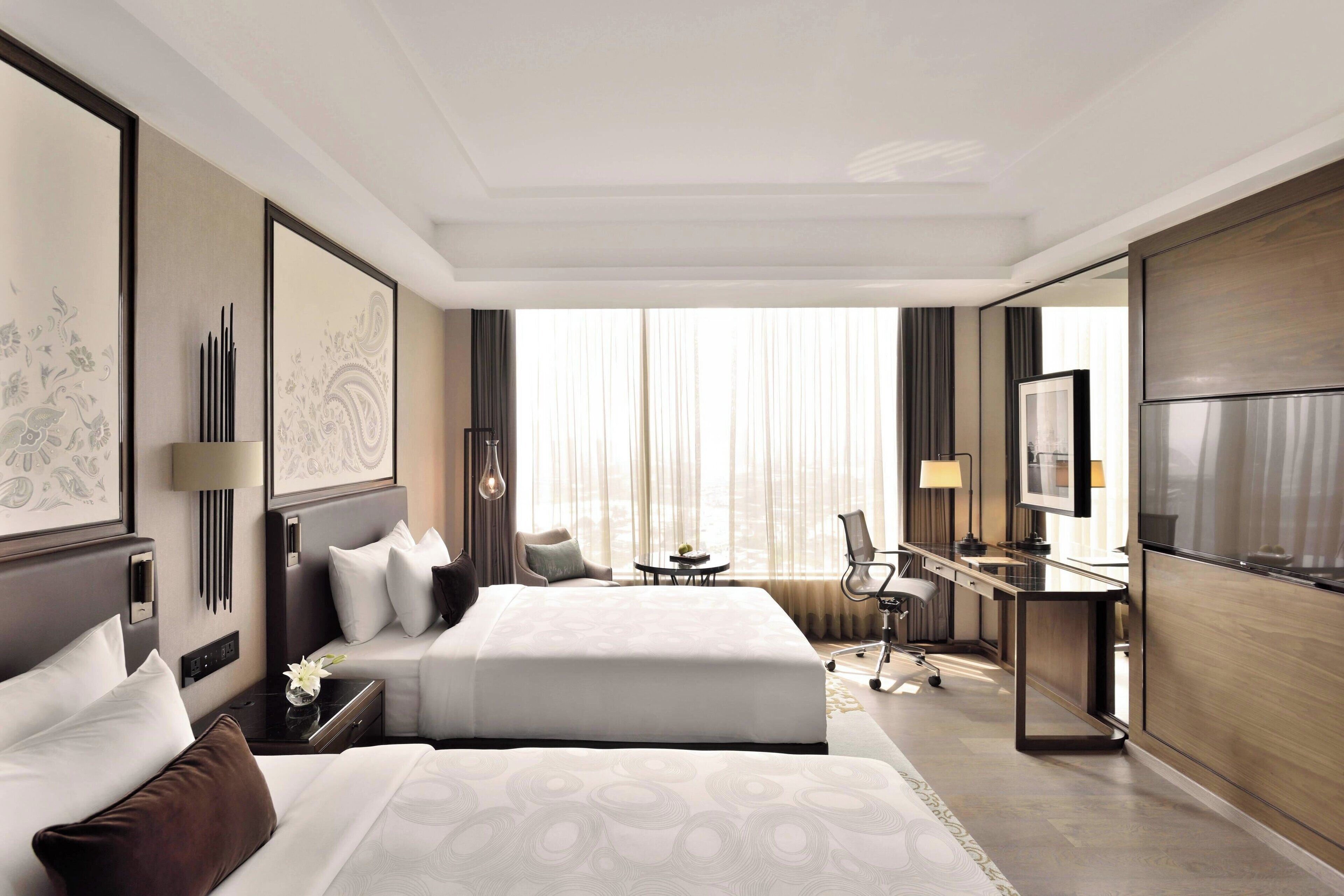 A hotel room with two beds, a desk, a chair, a TV, and a large window with curtains.