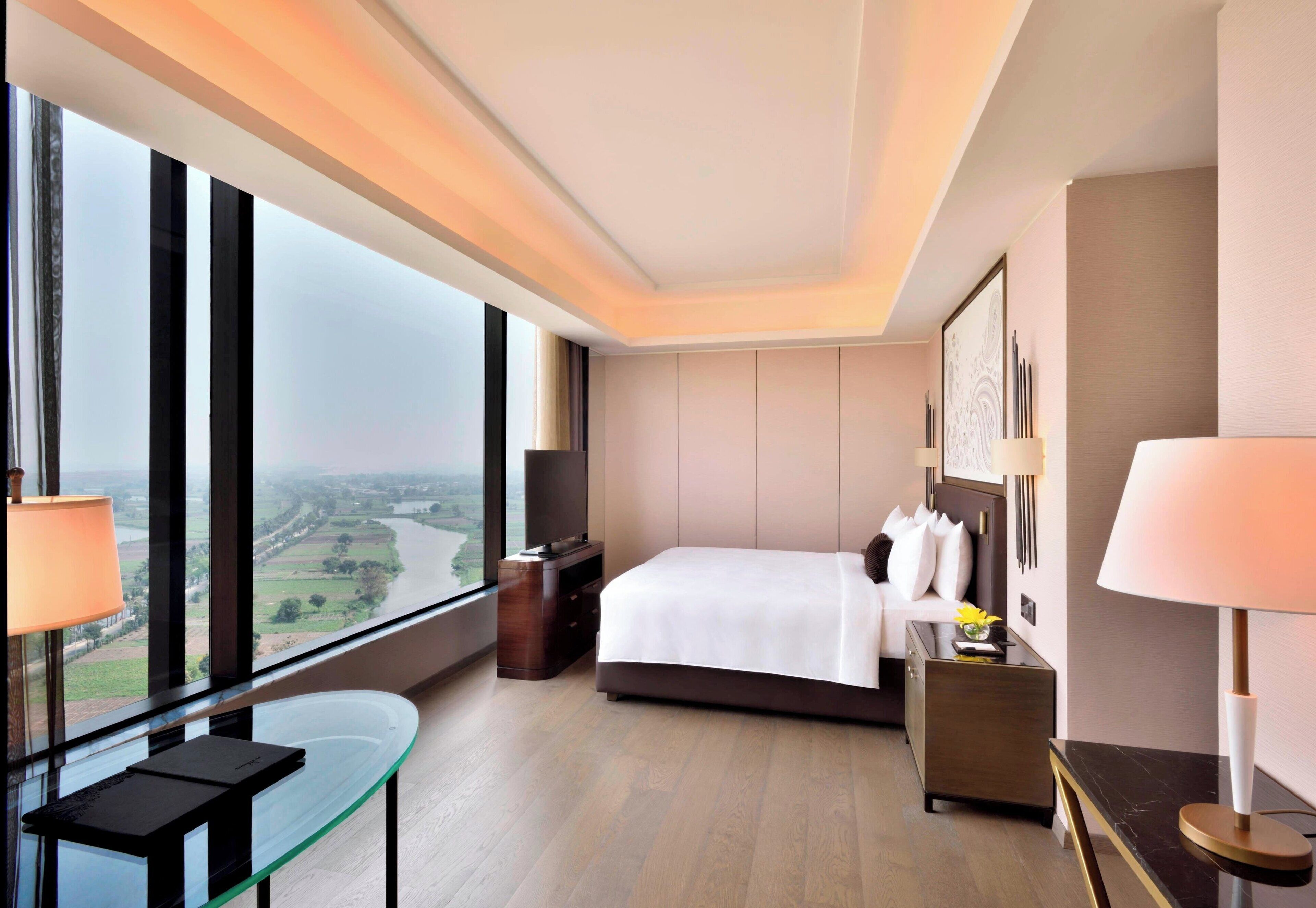 Habitación de hotel con cama, mesita de noche, lámpara, televisor y vistas al río a través de grandes ventanales.