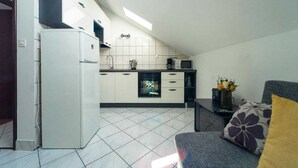 A compact kitchen with white cabinets, a refrigerator, and a microwave. There is a window providing natural light, and a small seating area with a sofa and a decorative pillow.